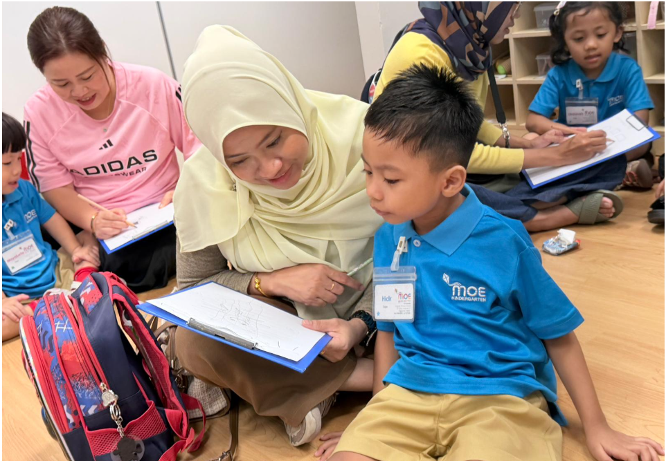 Woman in hijab helps a boy in blue uniform with a clipboard while others sit on the floor writing.